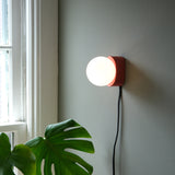 Round white globe light mounted on red square sconce with black cord hanging down on a gray wall near a window and large green leaf plant.