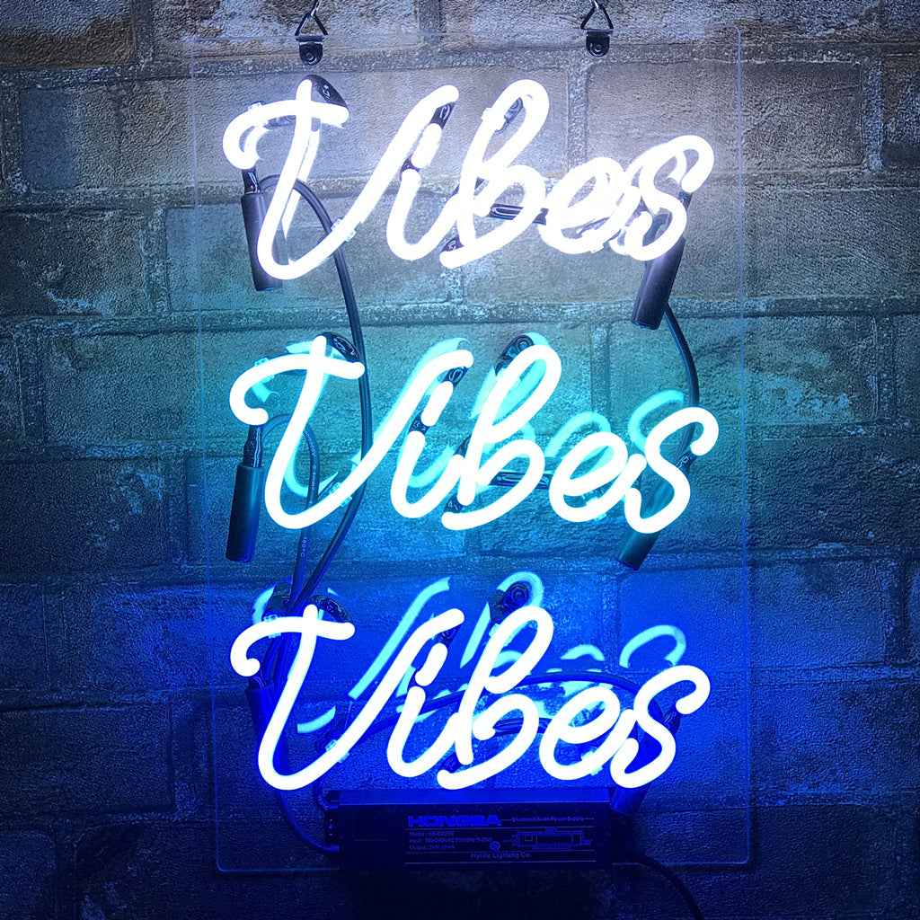 Three stacked neon signs displaying the word 'Vibes' in white, light blue, and dark blue script on a gray cement block wall.