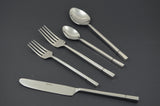 Five-piece stainless steel flatware set including a knife, two forks, a tablespoon, and a teaspoon with square-patterned handles on a dark background.