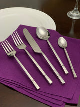 Five-piece silver stainless flatware set with geometric pattern handles arranged on a folded purple cloth napkin beside a white plate and wine glass on dark wood table.