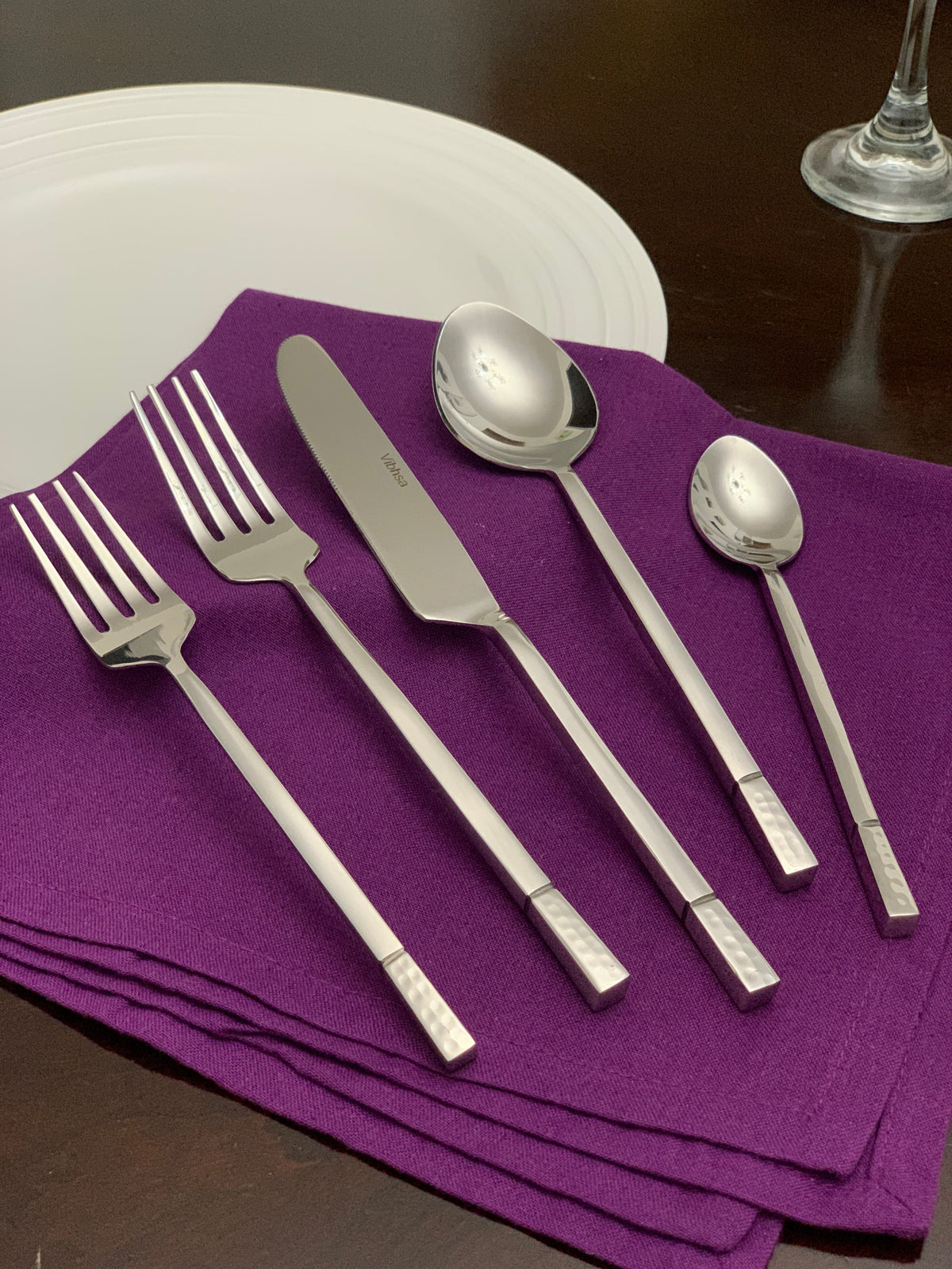 Five-piece silver stainless flatware set with geometric pattern handles arranged on a folded purple cloth napkin beside a white plate and wine glass on dark wood table.