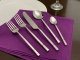 Five-piece modern stainless steel flatware set arranged on a folded purple cloth, including two forks, one knife, one tablespoon, and one teaspoon with patterned handles.