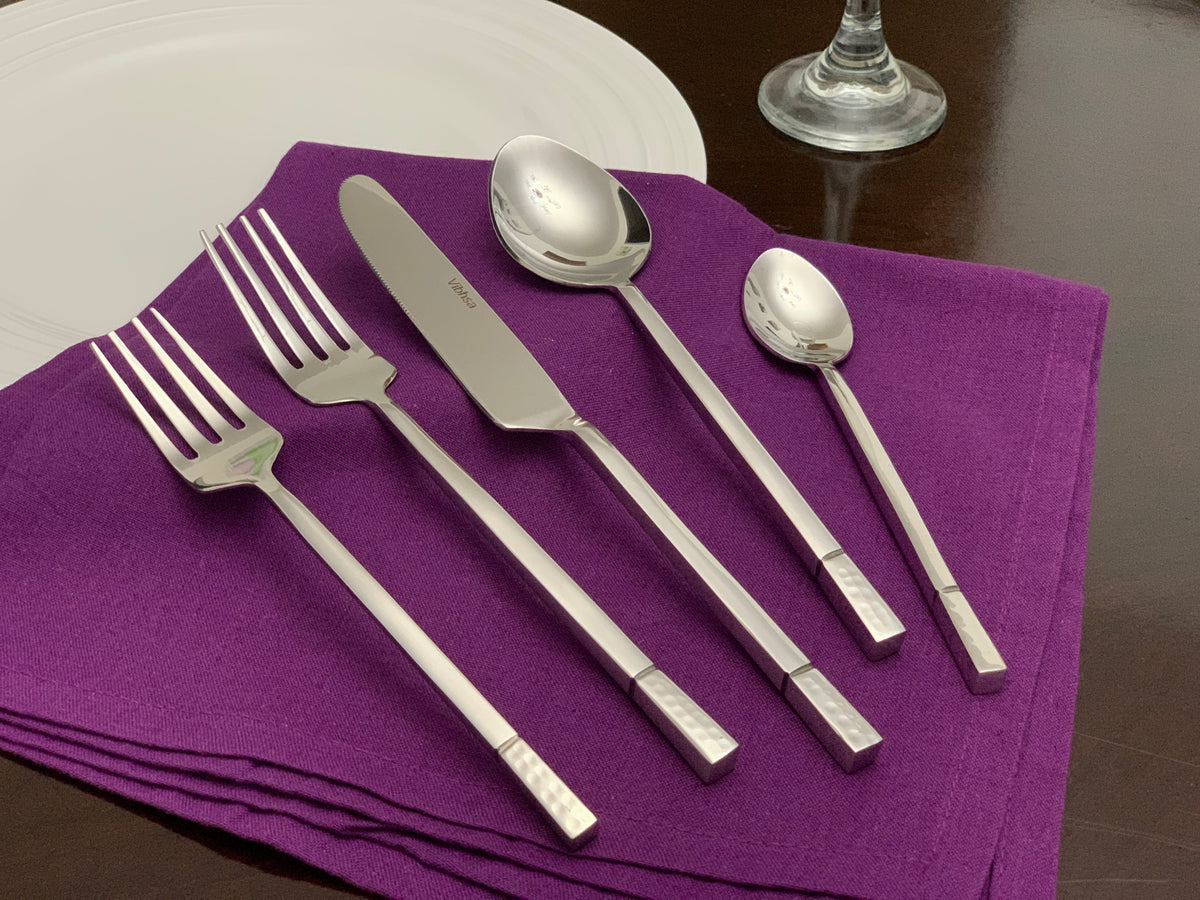 Five-piece modern stainless steel flatware set arranged on a folded purple cloth, including two forks, one knife, one tablespoon, and one teaspoon with patterned handles.