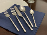 Set of five stainless steel flatware pieces with square patterned handles on a blue cloth napkin, including two forks, a knife, a soup spoon, and a teaspoon.