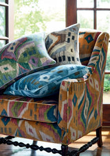 Upholstered armchair with multicolored abstract Ikat pattern in earthy tones, layered with velvet pillows in blue and muted geometric designs, beside a sunlit window.