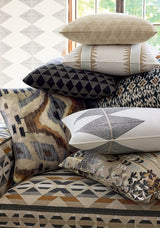 Stack of decorative pillows in beige, black, and cream with geometric and leaf patterns, arranged on a patterned fabric chair with earth tones.