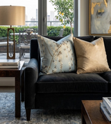Black velvet sofa with two large throw pillows, one light blue with a delicate floral vine pattern and the other solid beige, set next to a modern table lamp and window view.