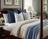 Bed with blue and cream patterned pillows, beige linen bedding, and a blue throw, framed by a dark wooden headboard and canopy posts in a softly lit room.