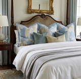 Bed with carved dark wood headboard and four blue pillows featuring white willow tree patterns, accented by smaller velvet and linen pillows, against pale curtains and a gold-framed mirror.