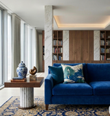 Blue velvet sofa with a patterned navy pillow featuring green willow tree design, set beside a wooden side table holding a blue-and-white ceramic vase and a small sculpture.