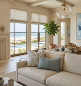 Neutral-toned living room with light beige sofa featuring a grey patterned pillow and a solid grey pillow, large windows with ocean view, and a glass pendant light.