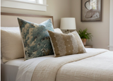 Bed with neutral beige bedding and two decorative pillows: one large in abstract blue and beige pattern, one smaller rectangular pillow with bronze intricate pattern.