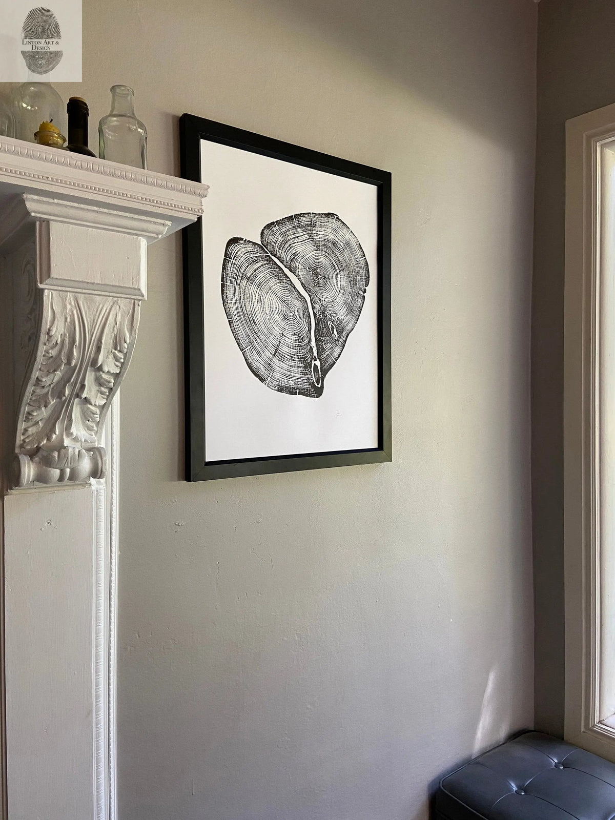 Black and white print of a cross-section of a cedar tree trunk, framed in black and hung on a beige wall next to a white fireplace and window.