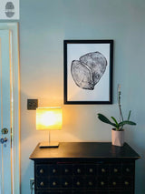 Black framed print of two overlapping tree rings hangs above a dark wooden cabinet with multiple drawers, illuminated by a nearby table lamp and accompanied by a potted orchid.