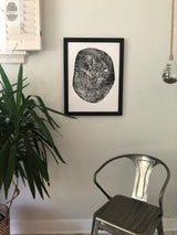 Black and white print of a pecan tree cross-section showing detailed growth rings, framed and hanging centered on a light wall above a silver metal chair and next to a green leafy plant.