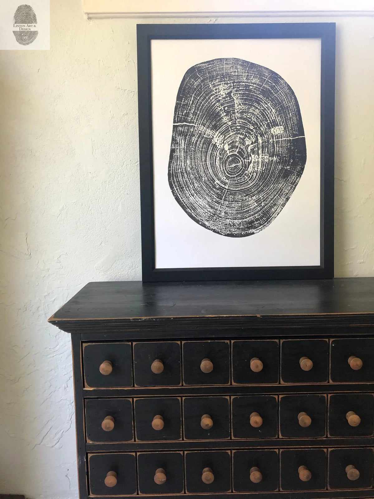 Black and white wood ring cross-section print framed in black, resting on top of a dark wooden chest with multiple small drawers and round knobs.