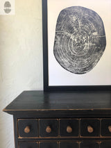 Black and white print of a pecan tree cross-section with visible growth rings, framed and leaning on a distressed black wooden dresser with five knobs.