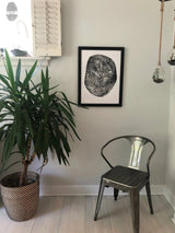 Black and white print of a pecan wood cross-section with visible growth rings, framed and hung on a light gray wall above a metal chair and beside a green leafy plant in a woven pot.