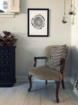 Black framed print of a detailed oak tree cross-section hung on a light gray wall above a beige upholstered wooden armchair with a black-and-white geometric pillow.