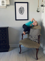 Black-framed print of a Massachusetts tree ring on a light gray wall above an antique-style chair with green upholstery and a black-and-white houndstooth pillow, with a child playfully perched on the chair's arm.
