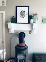 Black metal wood stove with red lid and pipe beneath white mantel holding framed tree ring print, green vase with plants, and glass bottle with pink ribbon on pale wall