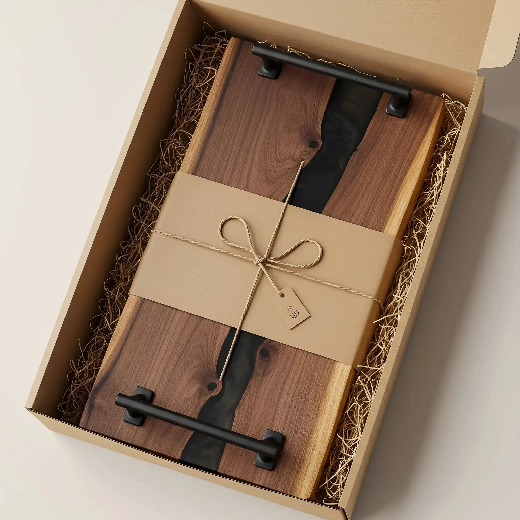 a charcuterie board carefully packaged for shipping. The board is made from live edge black walnut with a smooth, polished finish and a smoky black epoxy river running through the center. It’s wrapped in kraft paper and tied neatly with natural twine for a handmade, artisan feel. Matte black metal handles are attached at both ends.