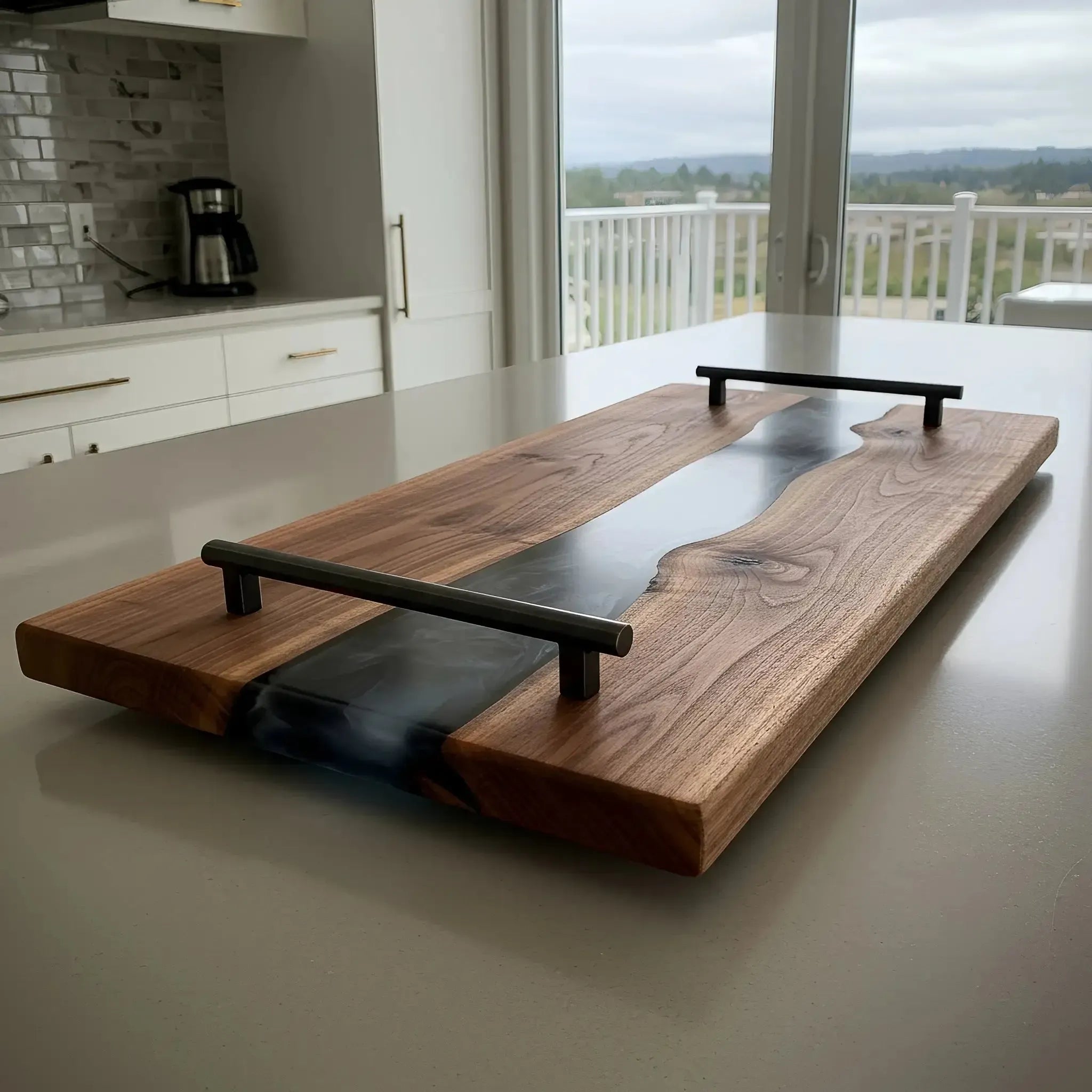 charcuterie board, 18 inches by 9 inches, made from live edge black walnut wood with a smoky black epoxy river running through the center. The resin has translucent depth, blending black, smoky gray, and hints of deep navy. The board has a polished walnut finish and matte black metal handles with a sleek design where the top bar slightly overhangs the vertical posts. The board is styled on a modern kitchen island and captured from a natural, slightly angled side or corner view.