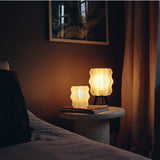 Two wavy-shaped lamps glowing warmly, one smaller and sitting on books on a textured round side table, the other larger on a tripod base beside beige curtains.