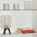 Two wavy glass lamps with light beige vertical ripples, one small cylindrical on a glass shelf and a larger one with black tripod legs on a white shelf beneath, next to stacked books.