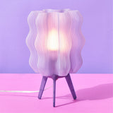 Purple wavy lamp with white inner light diffuser on three dark purple tripod legs against a pink and lavender background