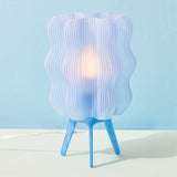 Table lamp with translucent white wavy shade and blue tripod legs, glowing softly against a pale blue background.
