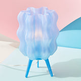 Translucent blue wavy lampshade with vertical ridges sitting on three slender blue legs, casting a soft light against pastel pink and blue backgrounds.