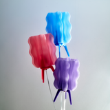 Three wavy, translucent lampshades in blue, pink, and purple, each supported by tripod-style legs matching their colors, arranged in a vertical cluster.