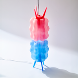 Vertical wavy lamp with stacked pink and blue translucent shades and blue legs casting a shadow on a white surface against a pale background.