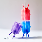 Three wavy, ribbed lamps in pink, purple, and blue with tripod legs, one hanging and two standing on a white surface against a plain light background.