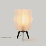 Table lamp with a rippled, translucent white shade shaped like overlapping waves, supported by three slender, black tripod legs on a light gray background.