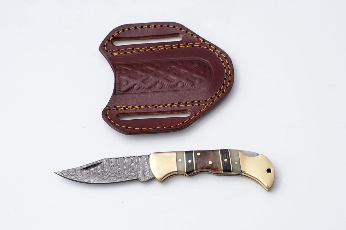 Brass & Bone Damascus Folding Knife with patterned steel blade and leather sheath laid on dark cloth