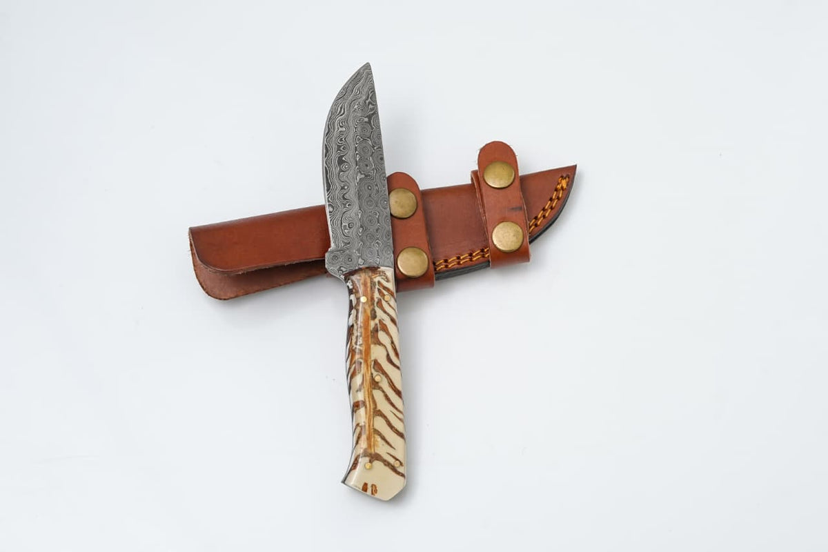 Handmade Damascus steel knife with camel bone handle and leather sheath resting on wood