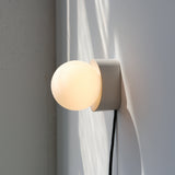 Wall-mounted sconce with a matte beige circular base and a glowing round frosted glass bulb, casting soft shadows on a light gray wall.