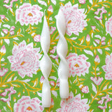 Two white twisted taper candles with smooth curves placed vertically on a green fabric featuring pink and white floral patterns with yellow stems.