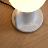 Close-up of a white ceramic or glass lamp base with a glowing spherical bulb on a light wood surface, featuring smooth glossy curves and a visible black power cord.