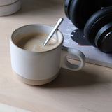 Matte ceramic coffee mug with a rounded handle featuring a finger hole, sitting on a light wood surface with a silver spoon inside and black headphones in the background.