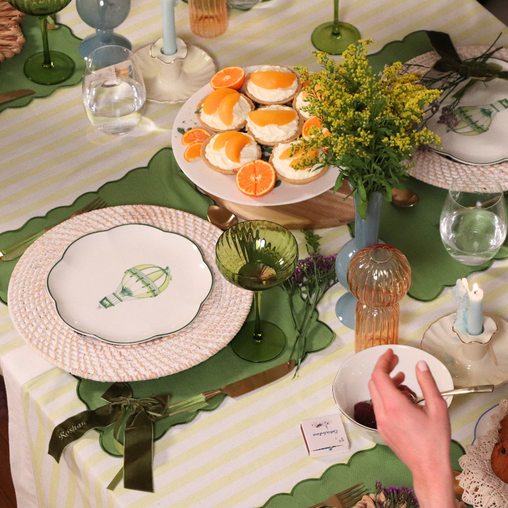 Table set with white plates featuring green hot air balloon designs on woven white rattan chargers, green scalloped placemats, and a centerpiece of desserts with orange slices and yellow flowers.