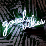 White neon sign spelling 'good vibes' in cursive mounted on a green textured wall with beige abstract patterns.