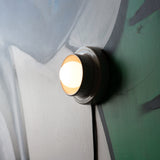 Round white wall sconce with a glowing bulb mounted on a multicolored wall featuring muted green, gray, and off-white brushstrokes.