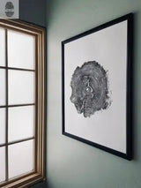 Wall clock shaped like a dark gray cross-section of an oak tree trunk with visible growth rings and light gray clock hands, mounted in a black frame on a muted green wall beside a window.