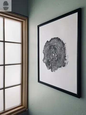 Wall clock shaped like a dark gray cross-section of an oak tree trunk with visible growth rings and light gray clock hands, mounted in a black frame on a muted green wall beside a window.