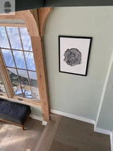 Black and white framed print of a detailed oak tree cross-section hangs on a pale green wall beside a large window with wooden trim in a room with light wood flooring and a dark tufted bench.