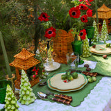Outdoor table set with green and white dishes, green glassware, woven wicker lanterns, fresh green vegetables, red flowers, and green, tree-shaped candles, creating a festive, natural centerpiece.