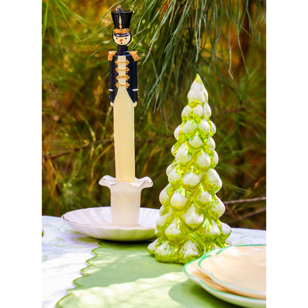 Green and white ceramic pine tree figurine with textured branches beside a nutcracker-themed candleholder on a green and white tablecloth with pine needles in background.